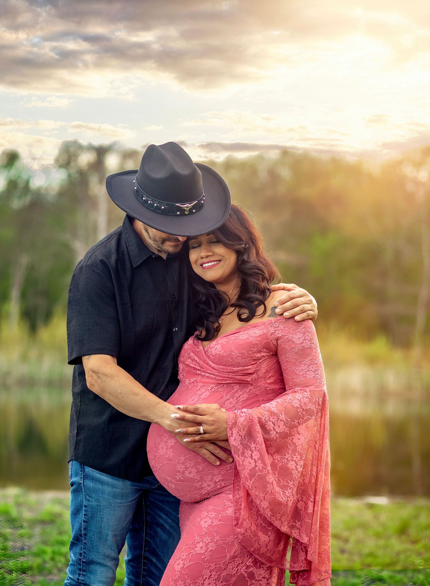 A couple poses outdoors at sunset, man in cowboy hat embracing pregnant woman in pink lace maternity gown.