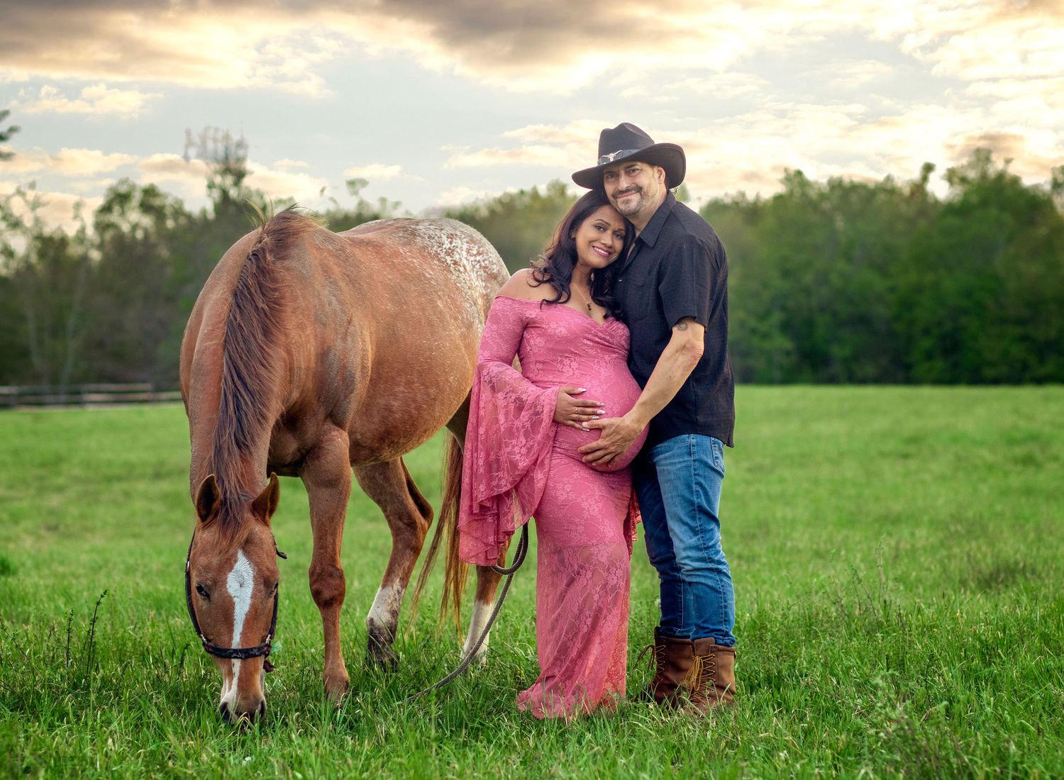 Pregnant couple in maternity photo with horse on green field, woman in pink dress, man in cowboy hat, sunset sky.