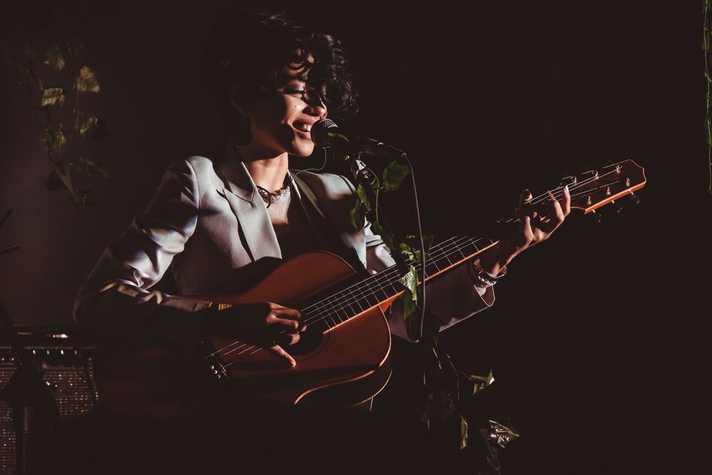Musician playing acoustic guitar and singing into microphone in a dark intimate venue.