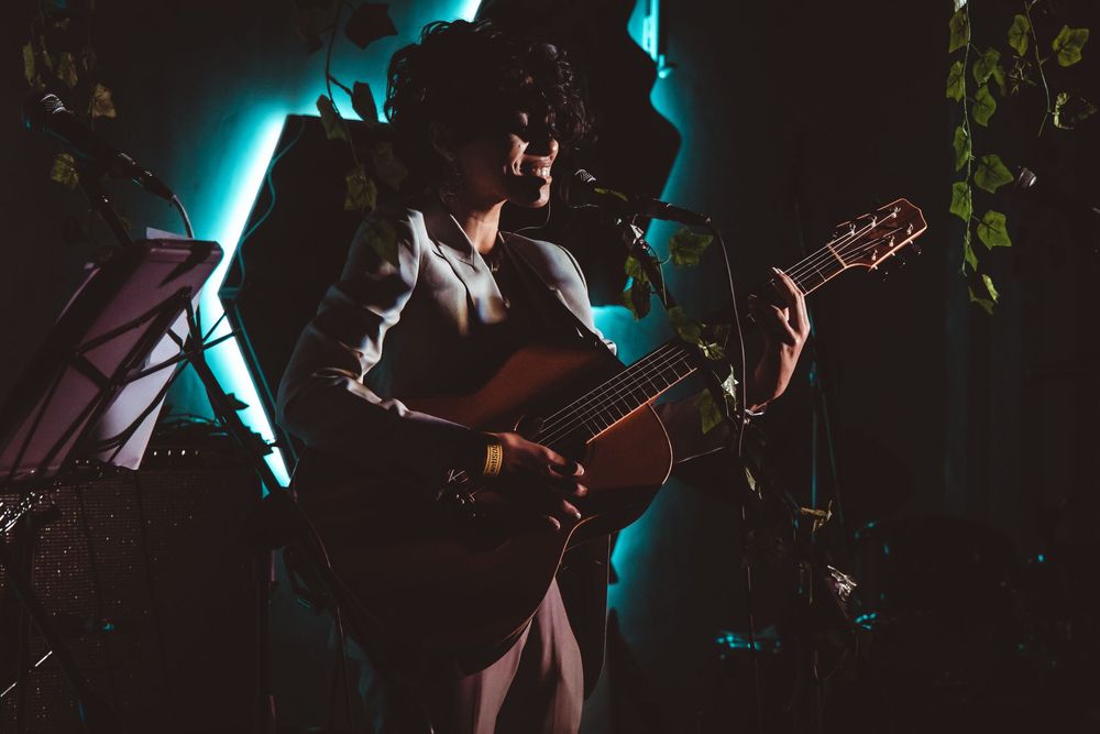 Female guitarist performing on stage under blue and purple stage lighting in a small venue.