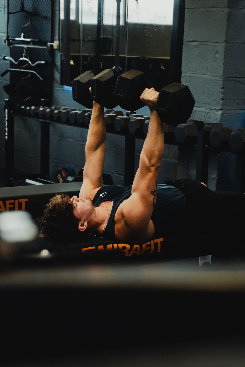 Muscular man performing dumbbell bench press exercise on an Emirafit bench in a dark gym.