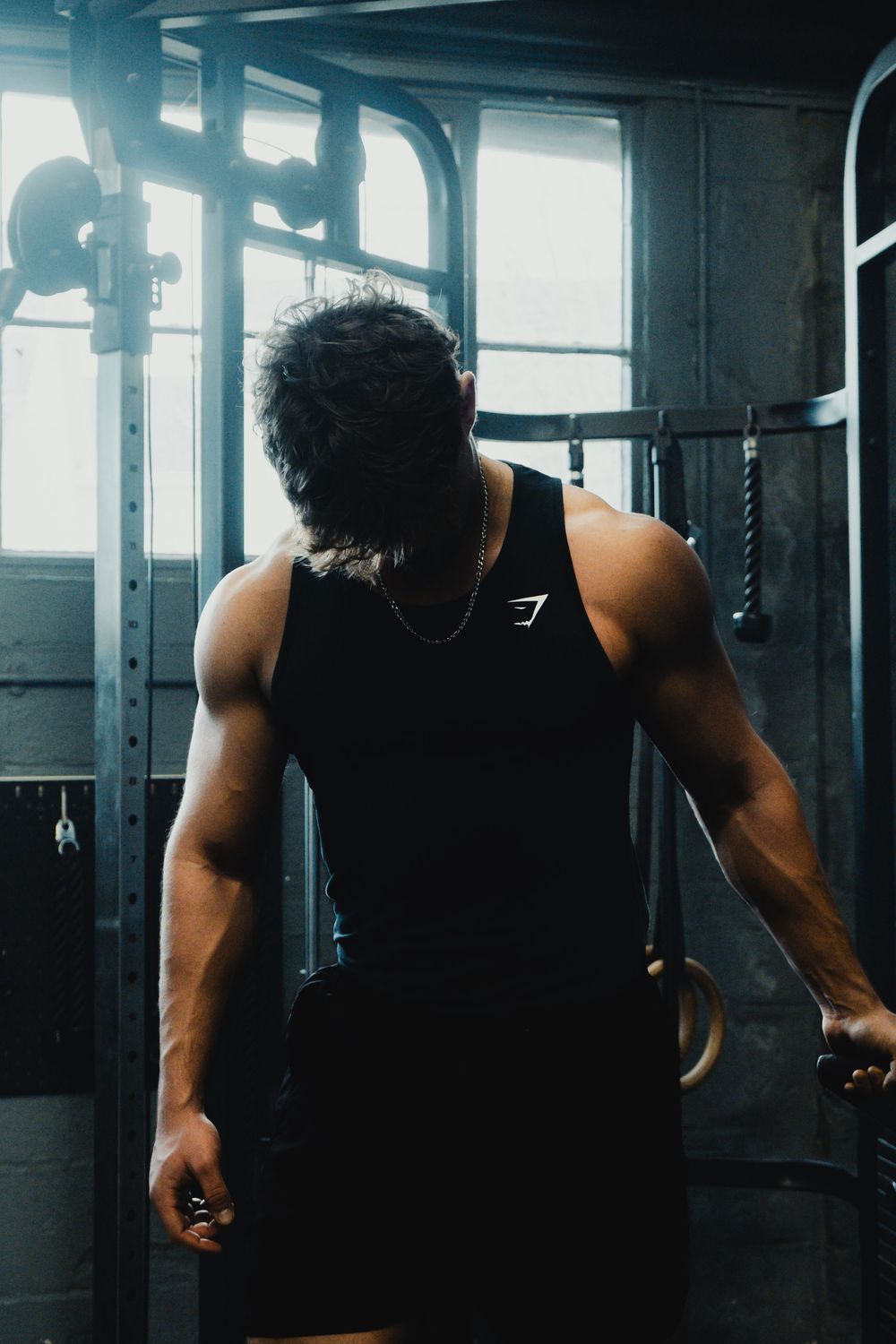 Muscular man in black Gymshark tank top working out on gymnastic rings in dark industrial gym.