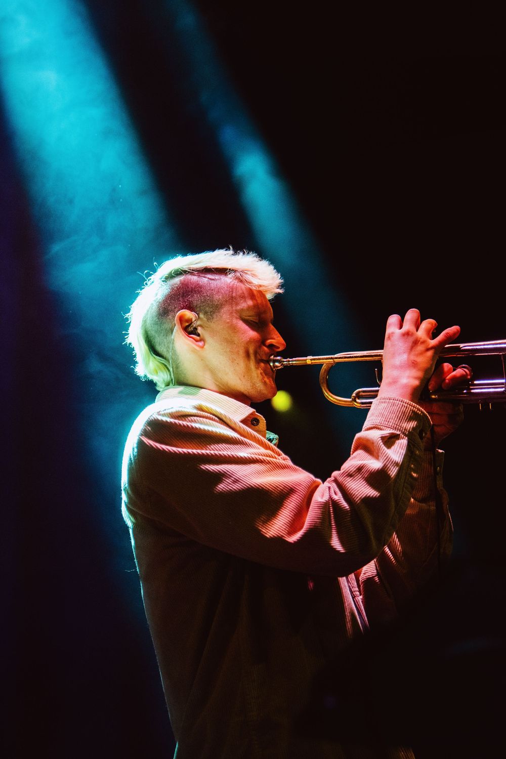 Musician with blonde hair plays trumpet on stage under dramatic blue and pink stage lighting.