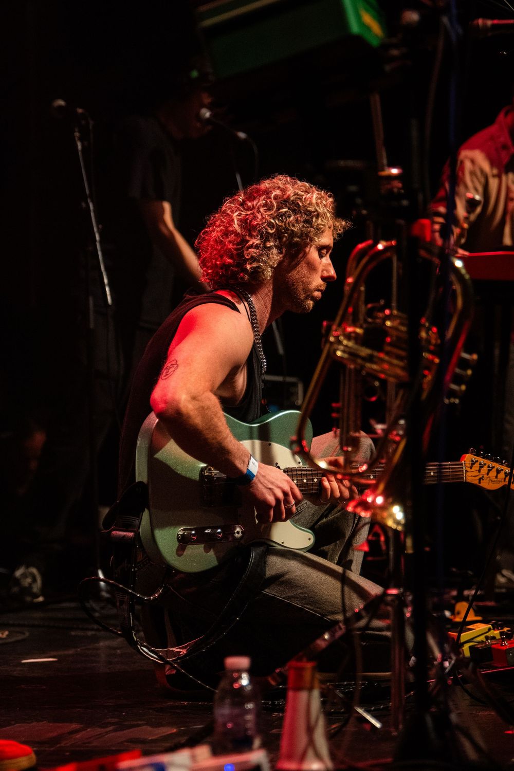 Curly-haired guitarist performs on stage under red lights, playing a green electric guitar at a live concert.