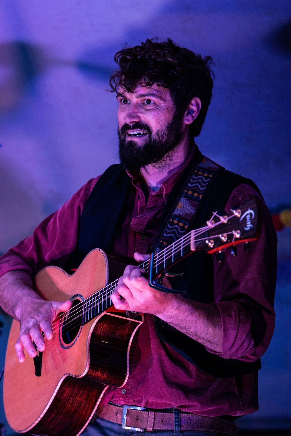 Bearded male musician performing live, playing acoustic guitar on stage under purple and blue lights.