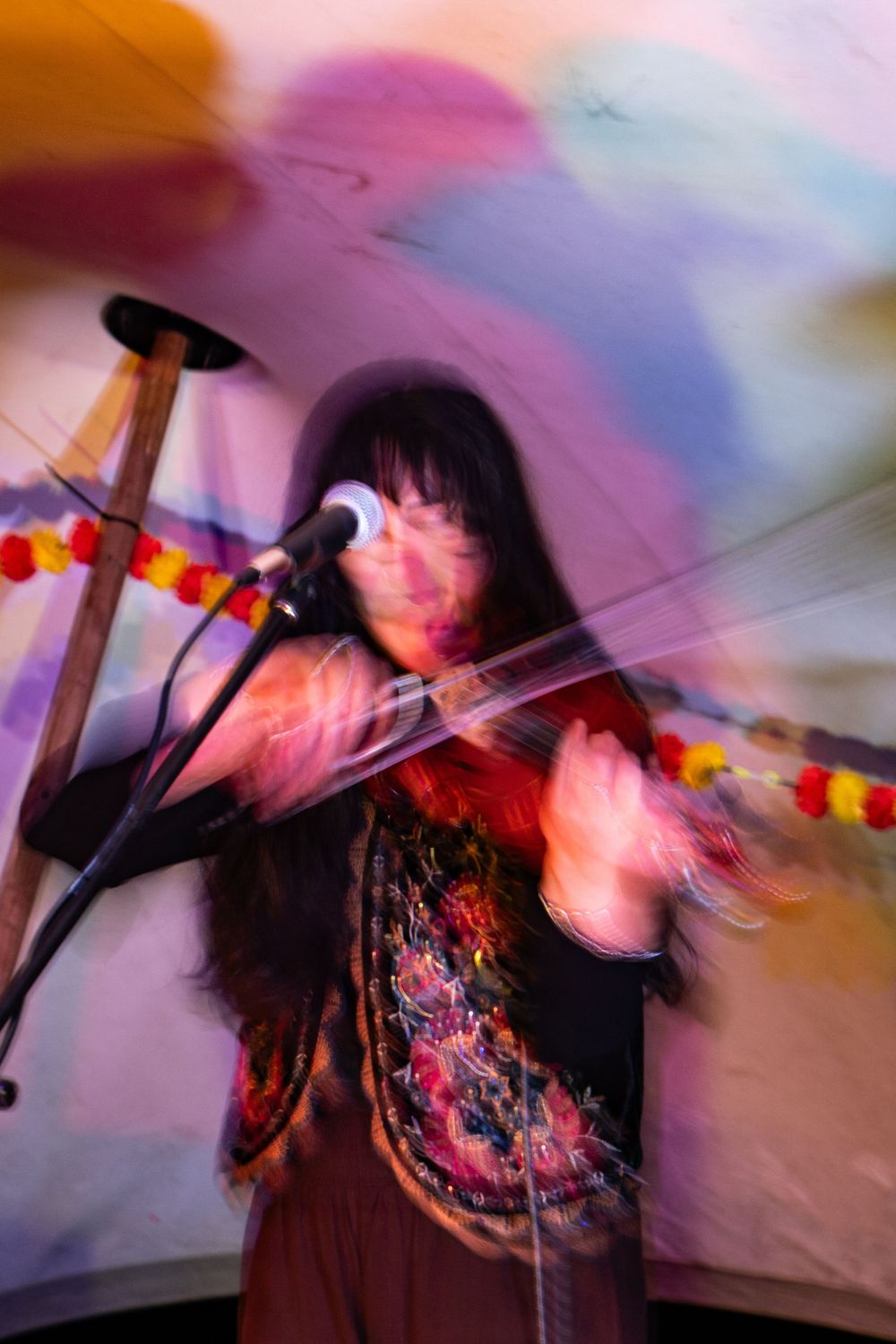 A woman with long dark hair plays violin at a microphone stand, captured with motion blur in a colorful venue.