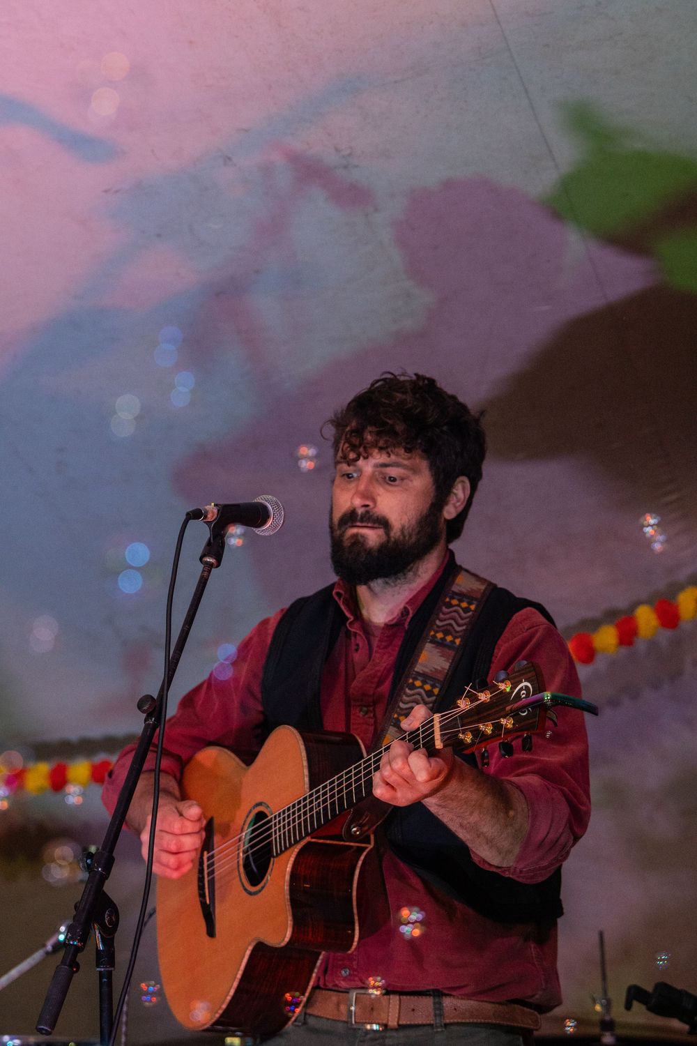 Bearded male musician performing acoustic guitar on stage at outdoor festival with microphone and colorful backdrop.