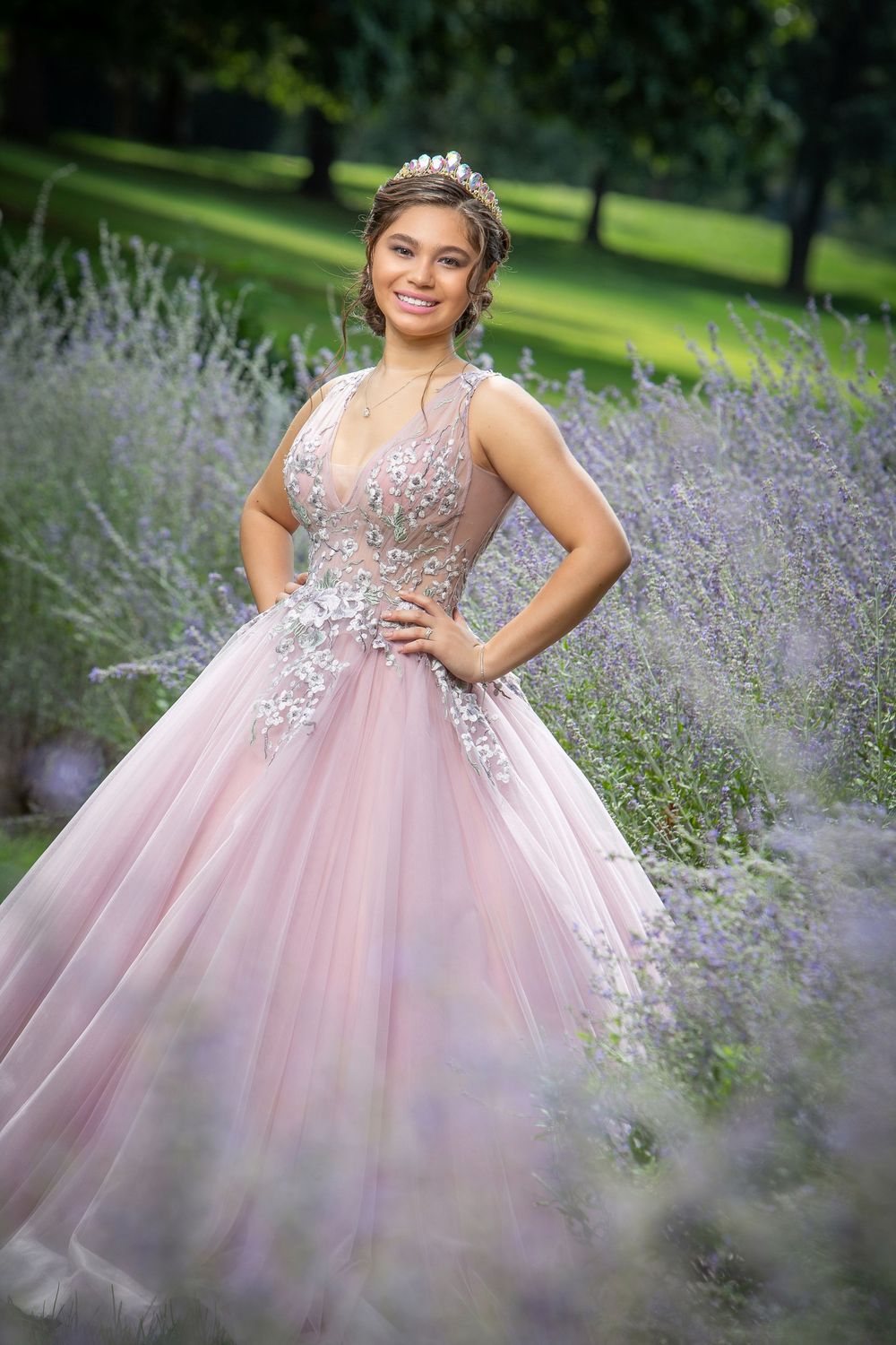 Young woman in pink floral ball gown and tiara poses gracefully among lavender blooms in a lush garden setting.