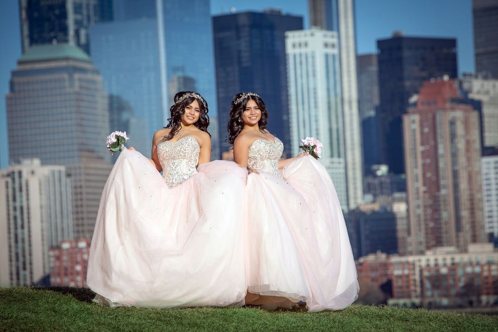 Two brides in blush ball gowns holding bouquets pose on a hill with a city skyline backdrop.