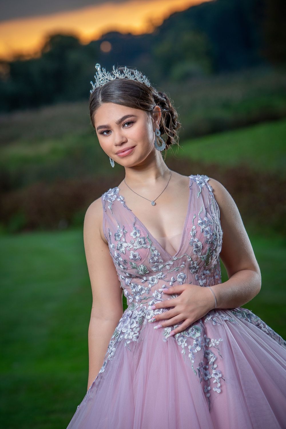 Young woman in pink floral ball gown and silver tiara poses outdoors at sunset for quinceañera or prom.