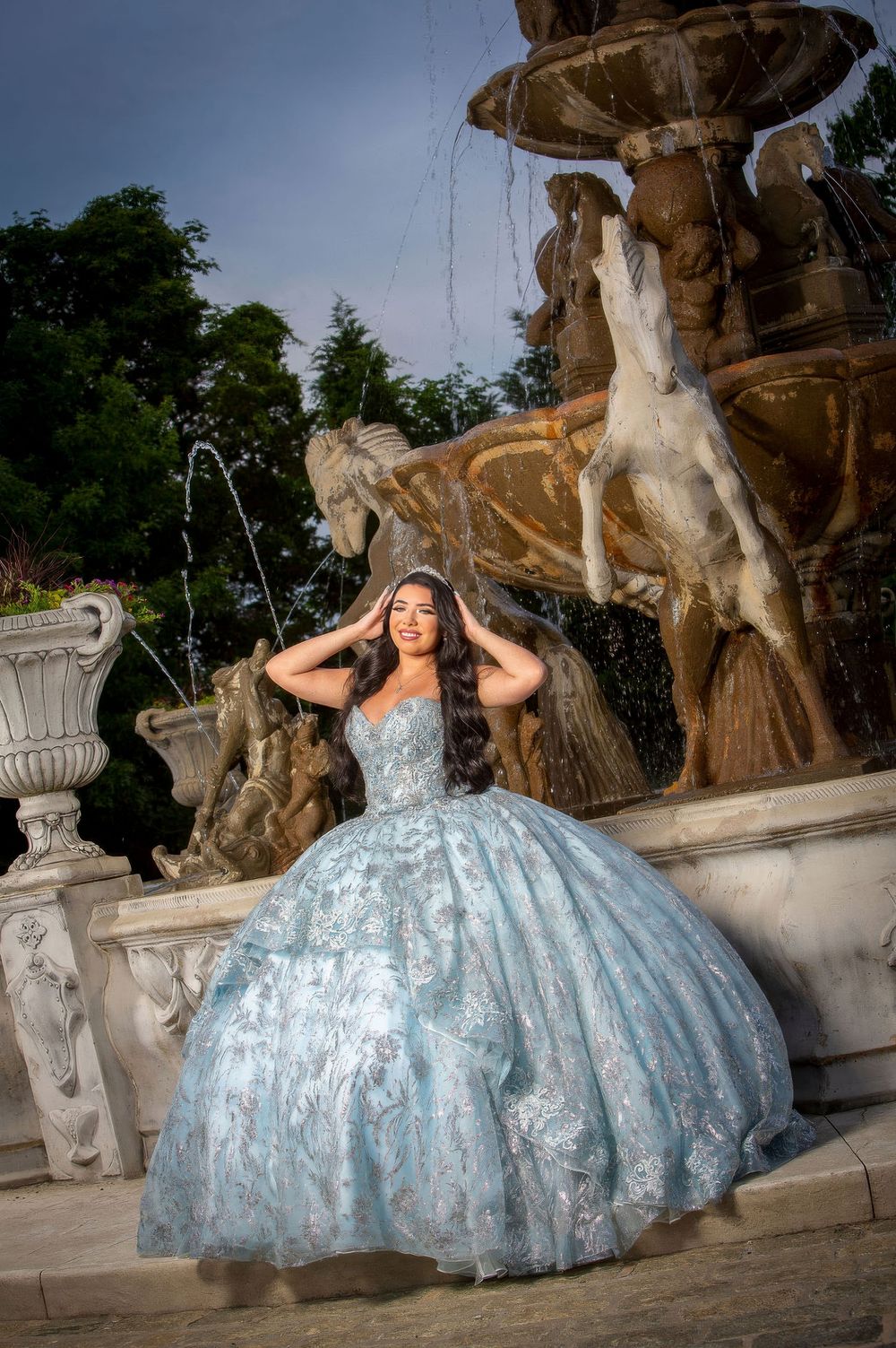 Young woman in light blue floral ball gown poses elegantly before ornate marble horse fountain outdoors.