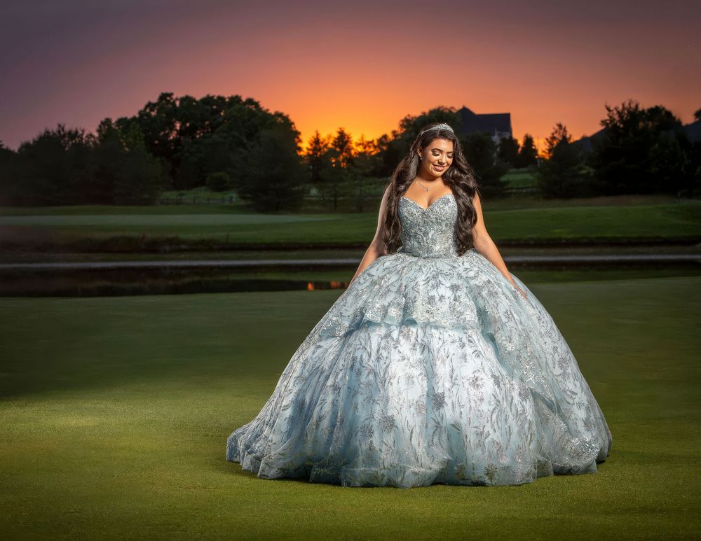 Young woman in light blue ballgown poses on golf course at sunset with vibrant orange sky.