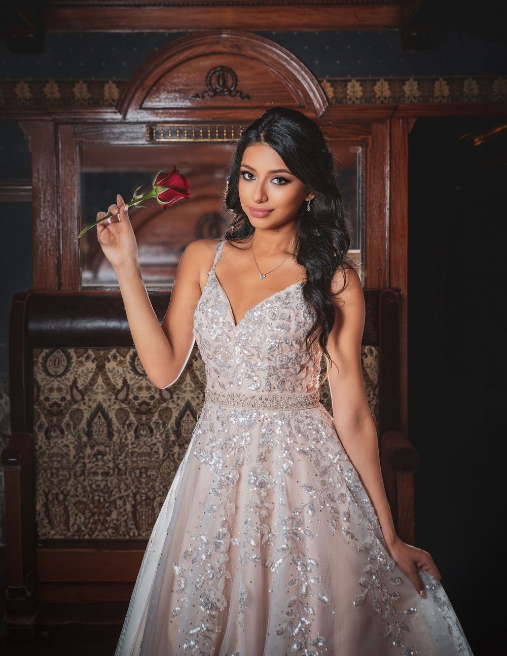 Young woman in elegant beaded ball gown holding a red rose, posing gracefully in a vintage-style interior setting.