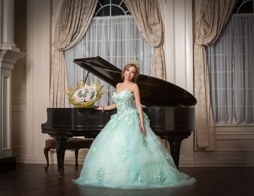 Young woman in mint green ball gown holding bouquet poses elegantly beside a black grand piano in ornate room.