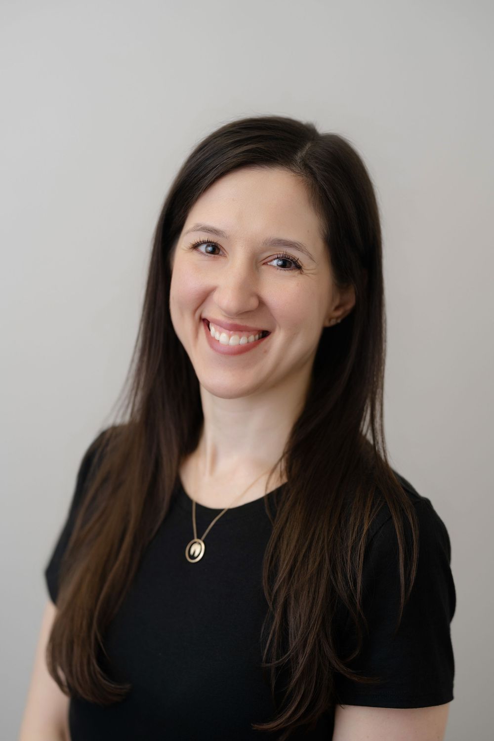 A smiling woman with long dark hair wearing a black top and gold necklace poses for a professional headshot.
