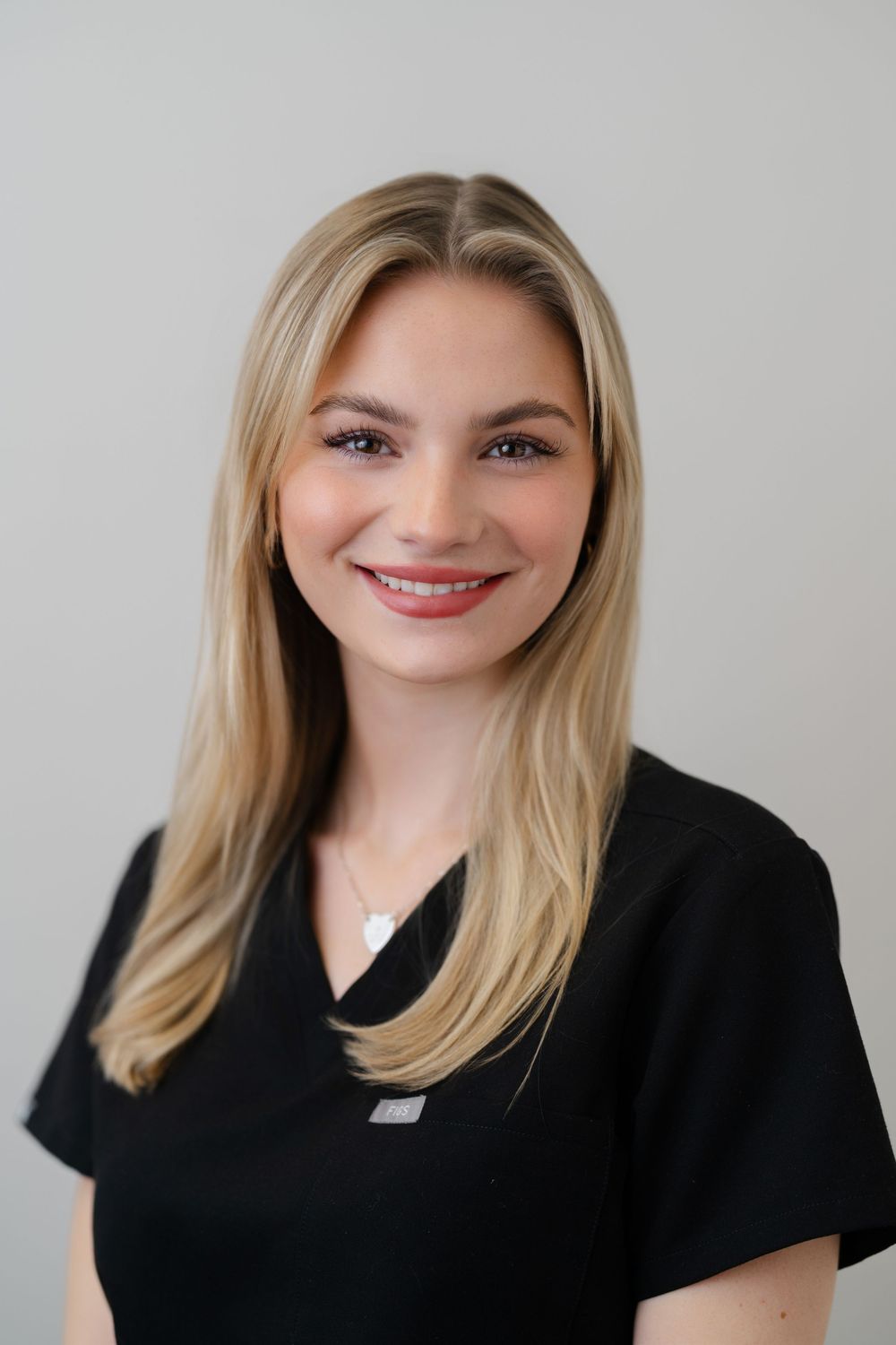 Smiling blonde woman in black scrubs, professional healthcare worker headshot on light gray background.