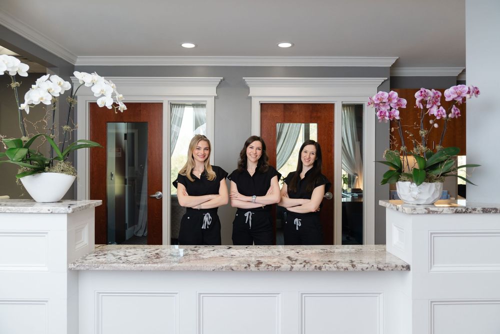 Three women in black uniforms stand with arms crossed behind a marble reception desk in an elegant white spa.