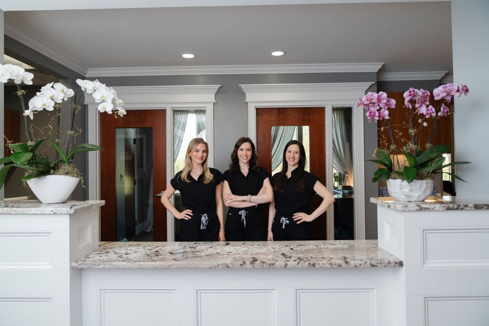 Three smiling women in black uniforms stand behind a marble reception desk in an elegant spa or salon lobby.