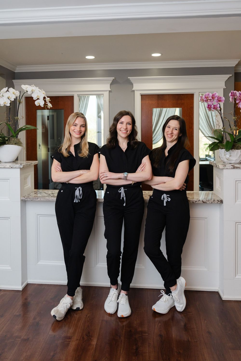 Three women in matching black scrubs stand with arms crossed in an elegant medical office reception area.