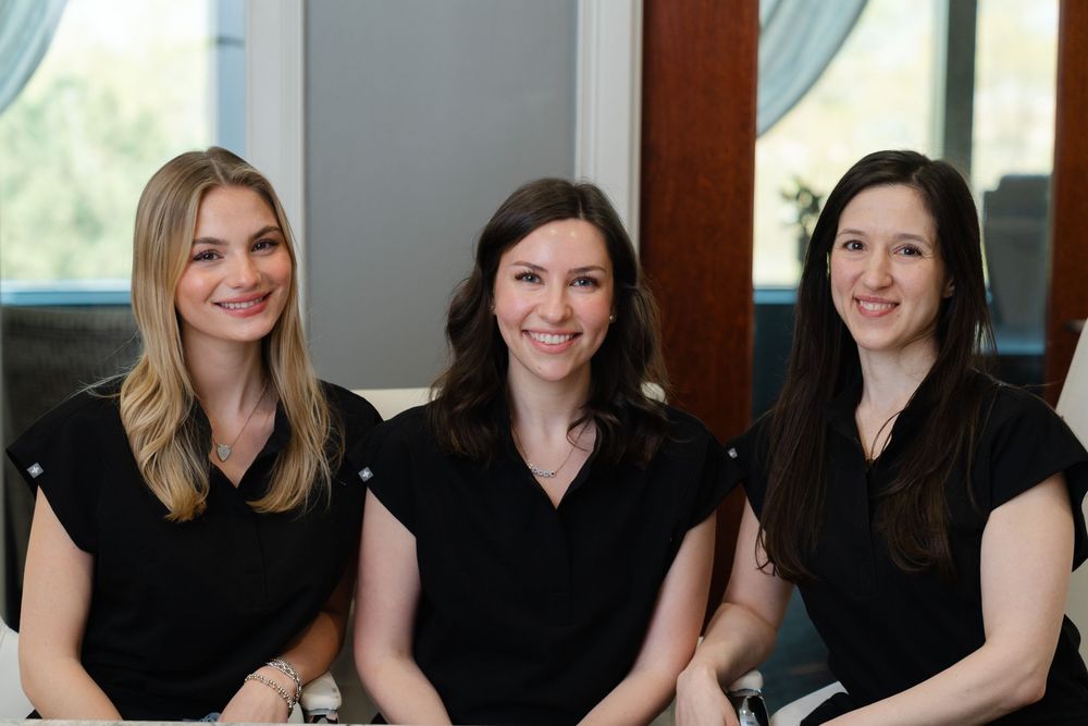 Three professional women in black tops smile while seated together in a bright modern office setting.