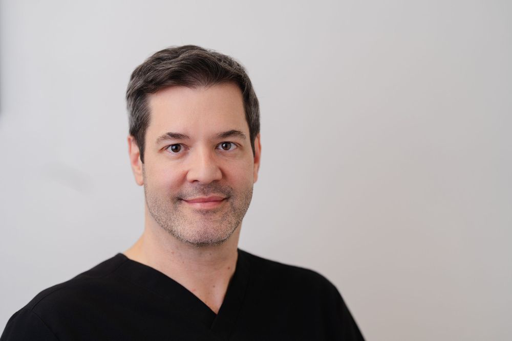 Professional headshot of a man with short dark hair wearing a black V-neck shirt against a light gray background.