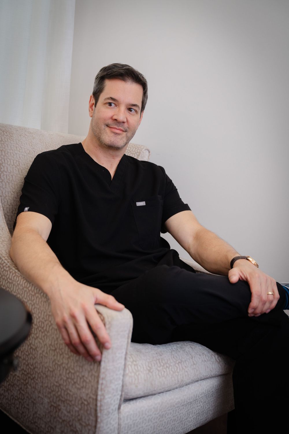 A male medical professional in black scrubs sits relaxed in a gray chair, smiling confidently in a modern clinic.