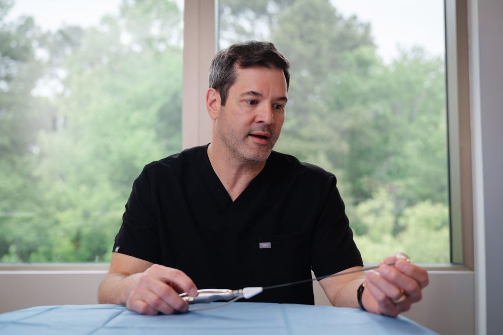 Medical professional in black scrubs holding surgical tools, seated at a table with trees visible through window behind him.