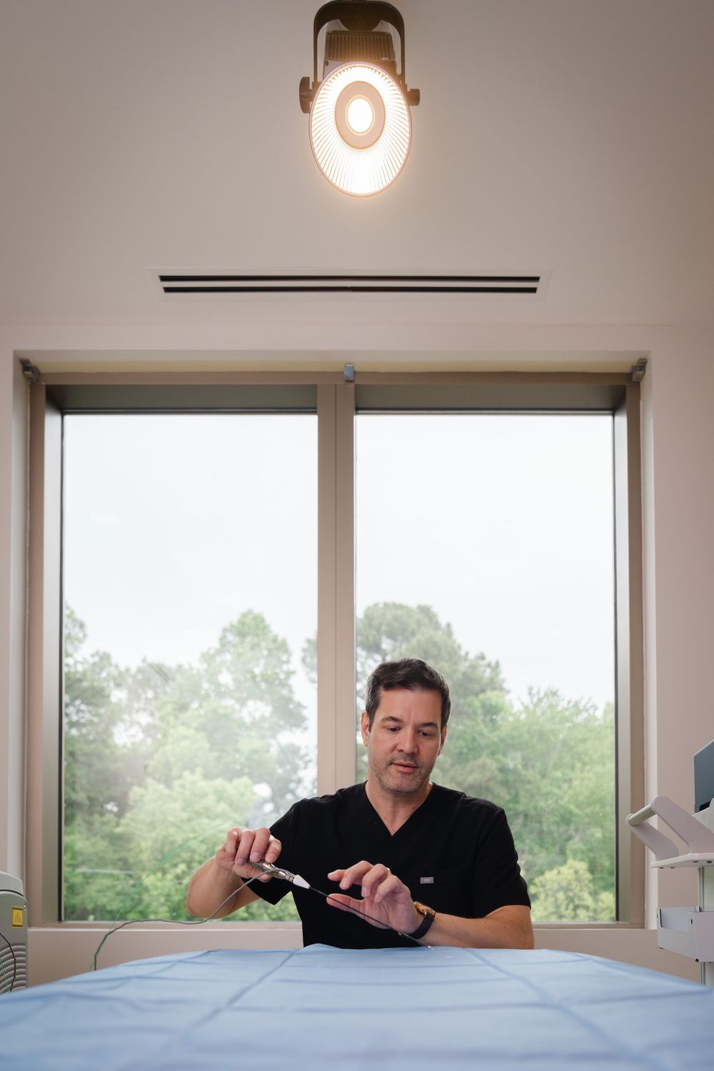 Medical professional in black scrubs holding surgical tools in a bright clinical examination room.