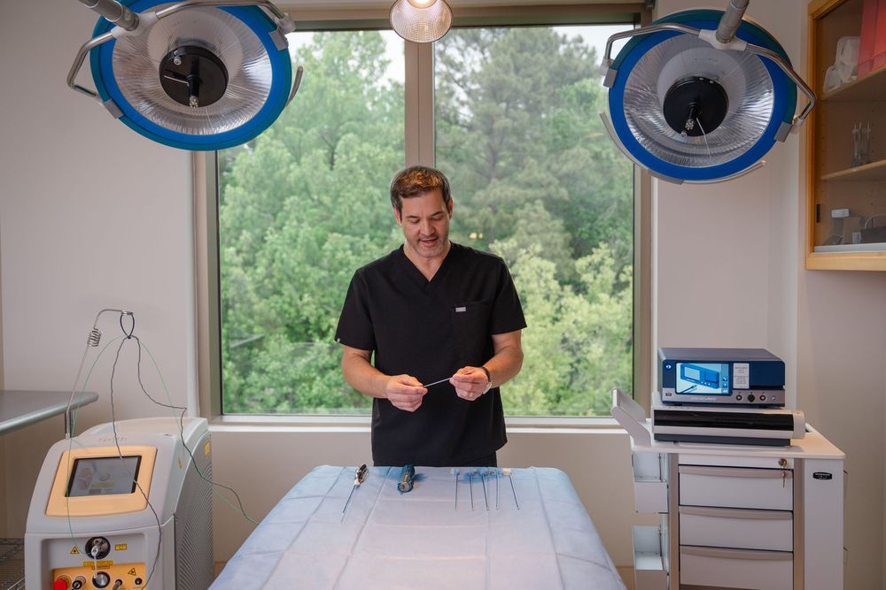 Medical professional in scrubs stands in a modern surgical suite with operating lights and medical equipment.