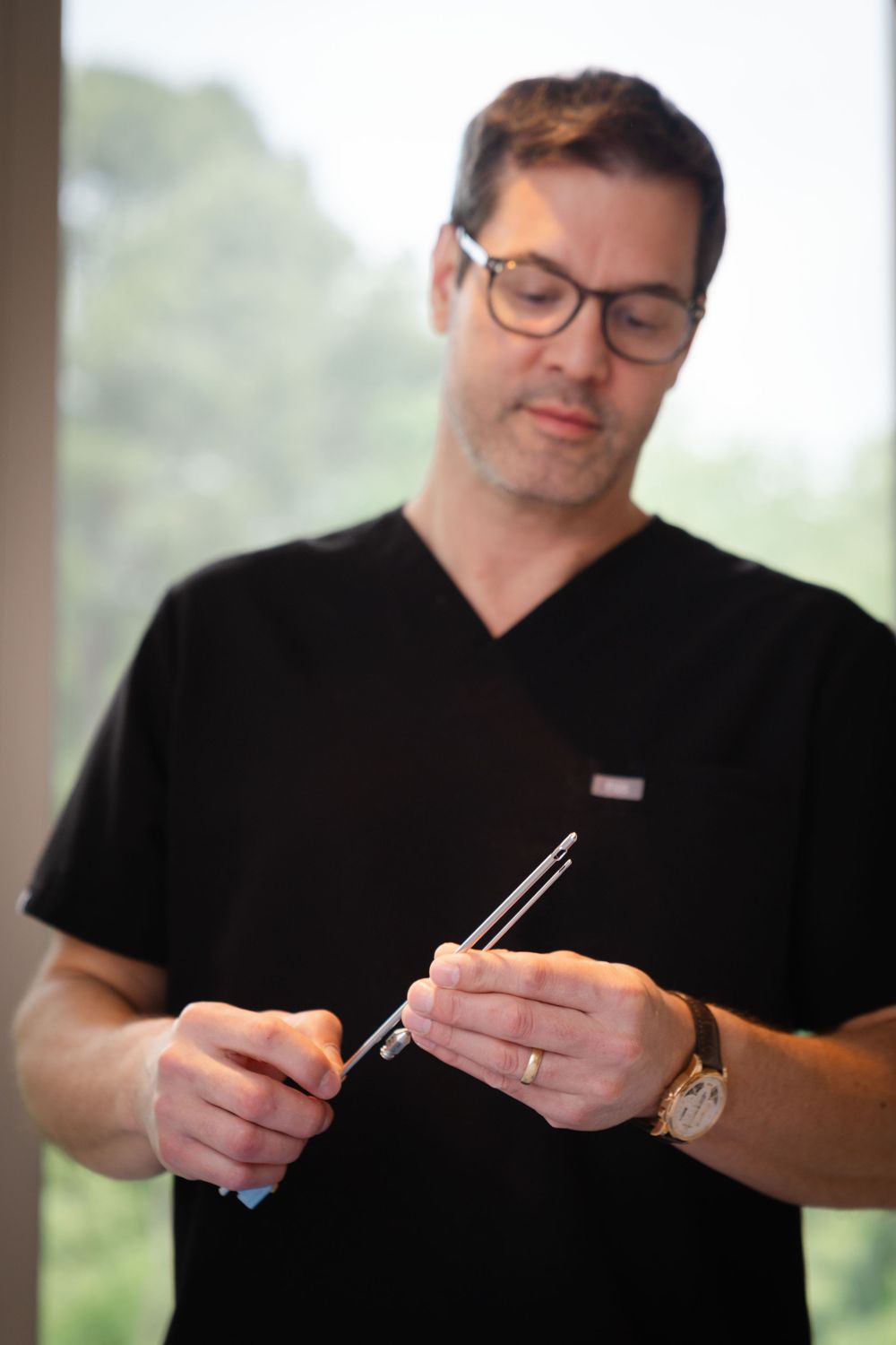 Doctor in black scrubs and glasses examining surgical instruments with a gold watch visible.
