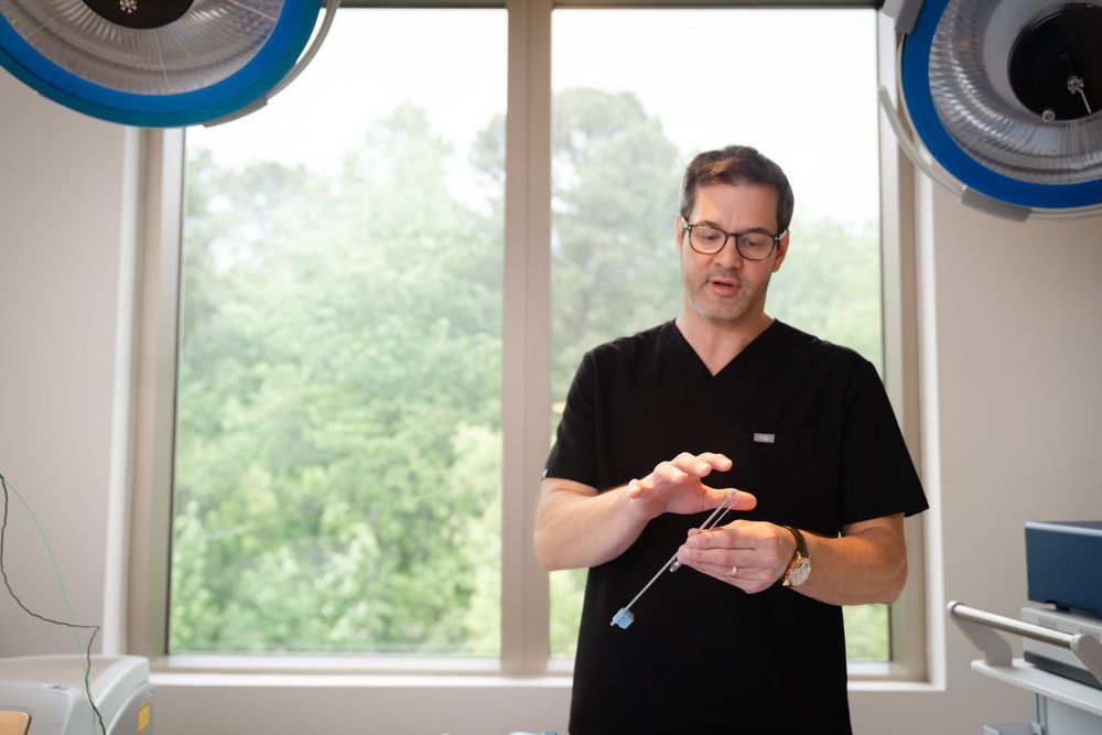 A surgeon in black scrubs examines a medical instrument in a bright clinical room with surgical lights.