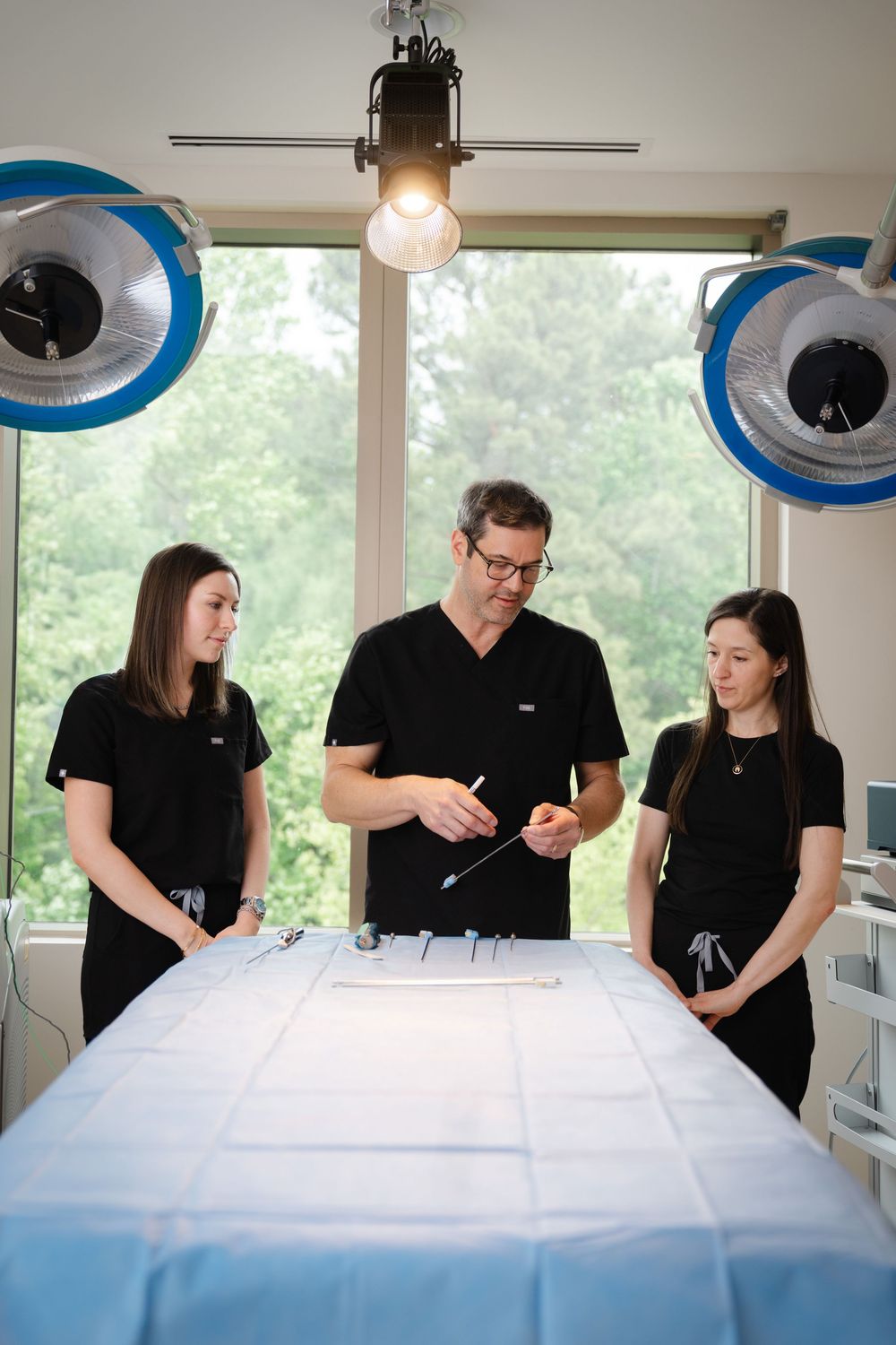A male surgeon in black scrubs instructs two female medical staff over a draped procedure table in a surgical suite.