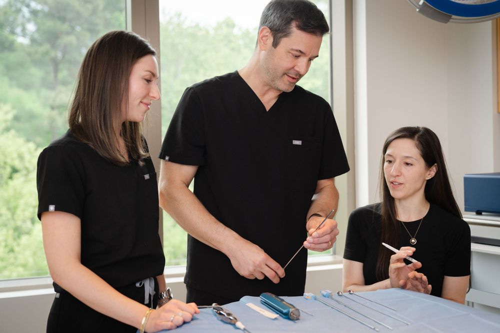 A male doctor in black scrubs reviews medical tools with two female colleagues at a clinical preparation table.