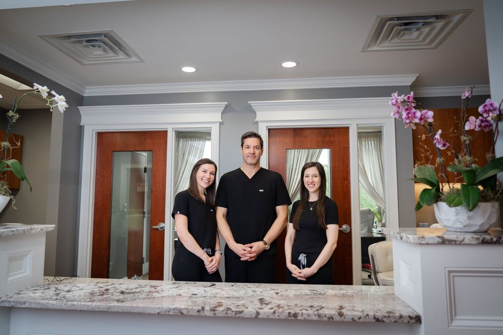 Three dental staff members in black scrubs smiling behind a marble reception desk in a modern dental office.