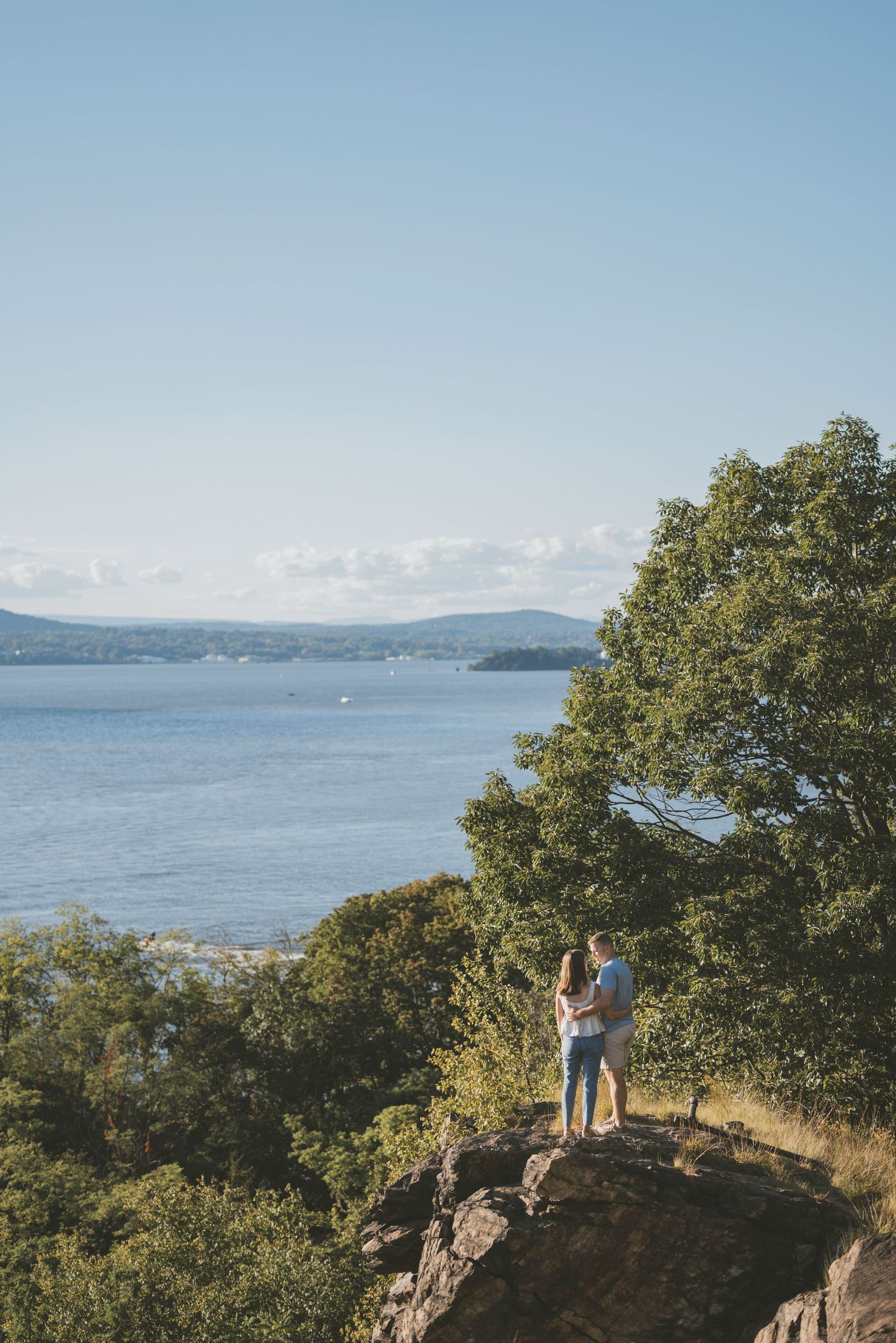 Couple embracing on rocky overlook surrounded by lush trees with scenic lake and distant hills under blue sky.