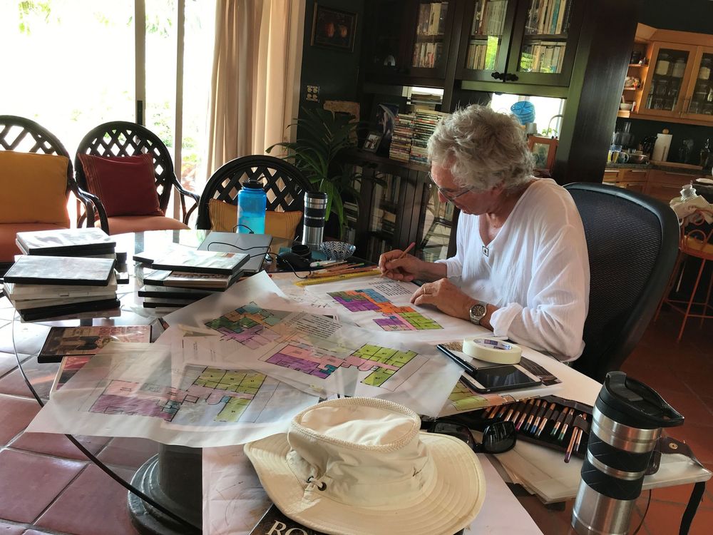 Woman with white hair sits at glass table covered in colorful maps and papers, writing in a bright sunlit room.