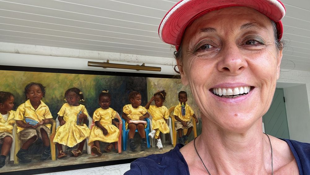 Smiling woman in red cap poses for selfie in front of colorful painting of children in yellow dresses.