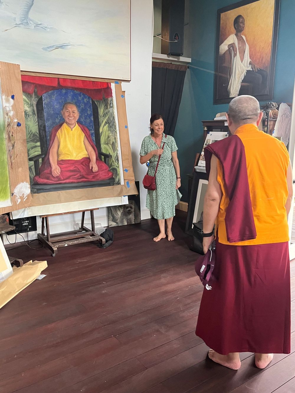 Buddhist monk in maroon robes views a colorful portrait painting of a lama on an easel in an art studio.