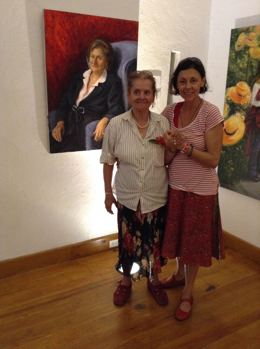 Two women posing together at an art gallery beside a painted portrait displayed on a white wall.