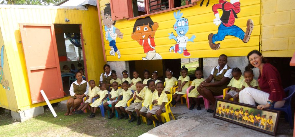 Children and teacher pose outside a colorful school building with cartoon murals in a tropical setting.