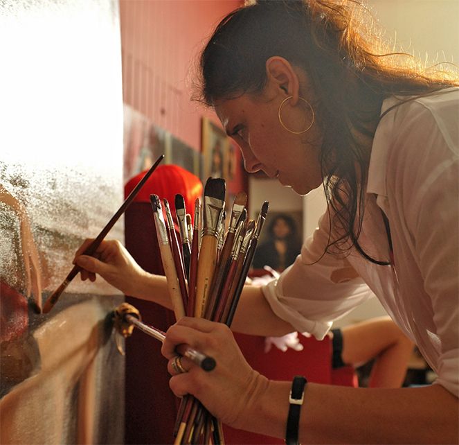 Young female artist holding multiple paintbrushes while working on a canvas painting in her art studio.