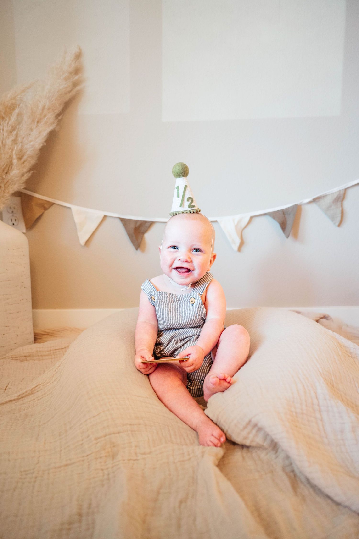 Smiling baby in gray overalls wearing a half birthday crown sits on beige cushions with bunting banner backdrop.
