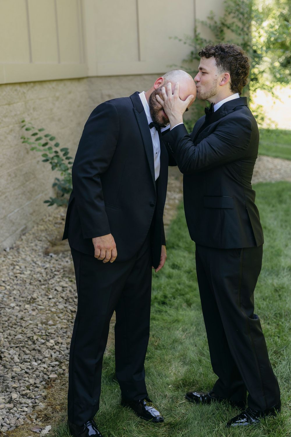 Two men in black tuxedos share a playful moment outdoors, one kissing the other's bald head at a   Wisconsin wedding day