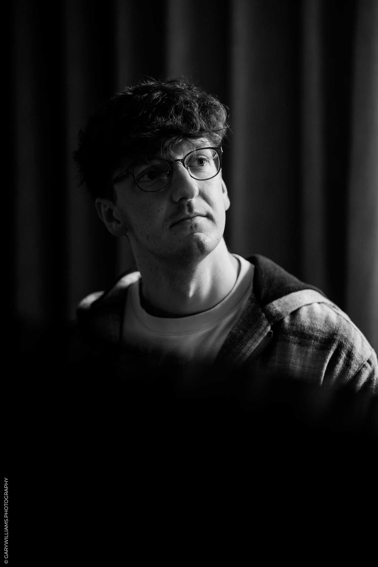 Black and white portrait of a young man with curly hair and glasses looking upward in dramatic lighting.
