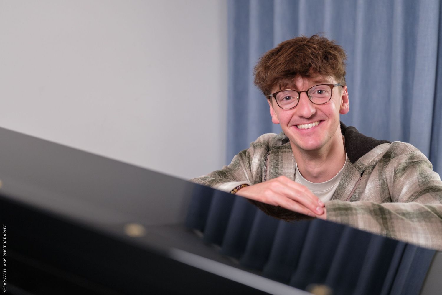 Smiling young man with glasses leaning on a black piano in front of blue curtains.