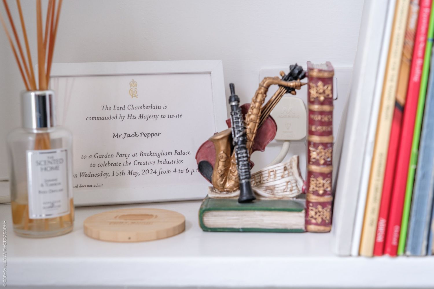 Shelf display with framed Buckingham Palace garden party invitation, saxophone figurine, vintage books, and reed diffuser.