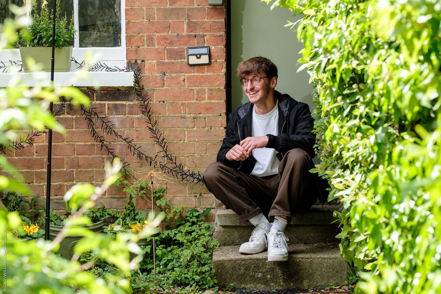 Young man smiling, sitting on stone steps outside a brick house surrounded by lush green garden foliage.