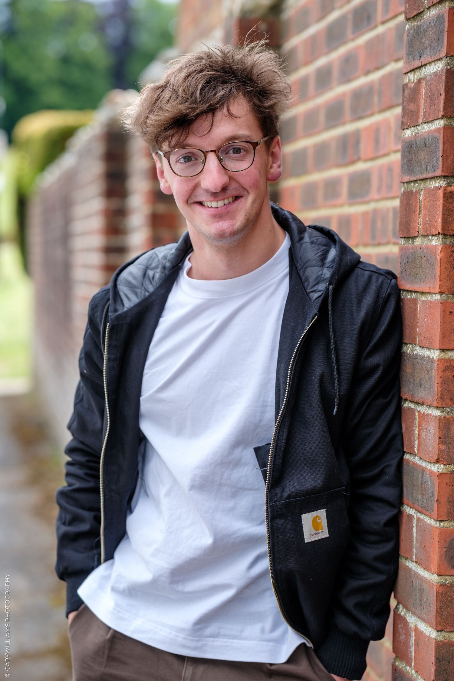 Young man with glasses smiling, wearing a Carhartt tee and black jacket, leaning against a brick wall outdoors.