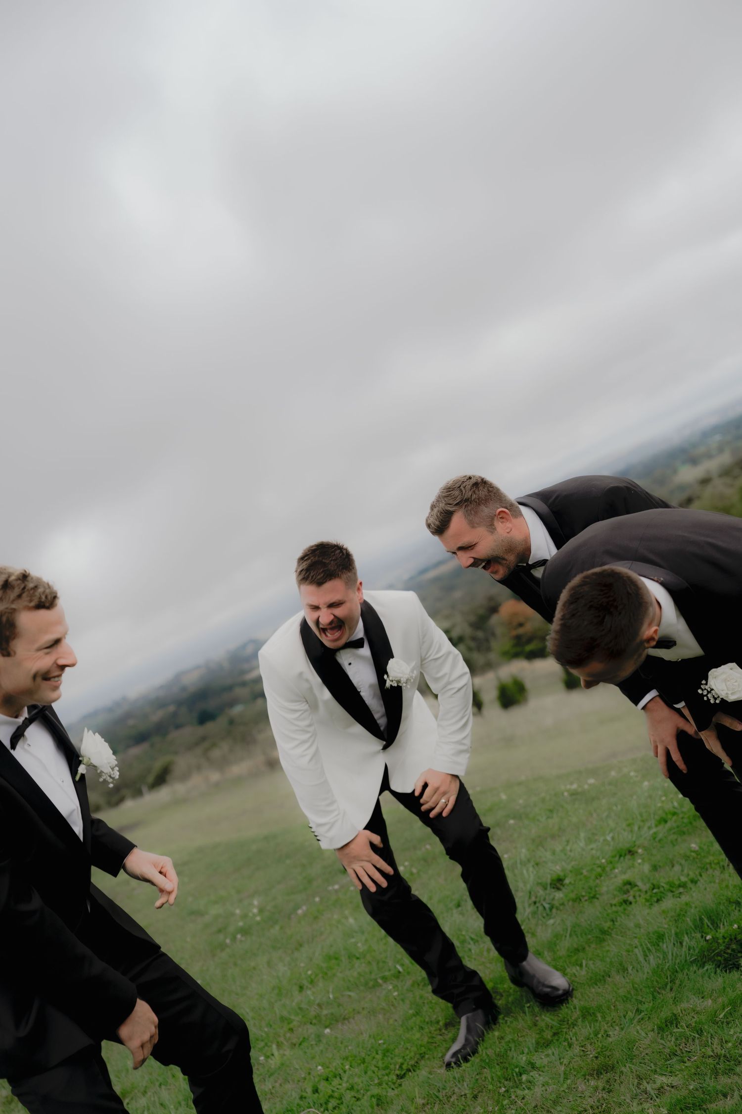 Groom in white tuxedo laughing with groomsmen in black suits on green hillside at outdoor wedding.