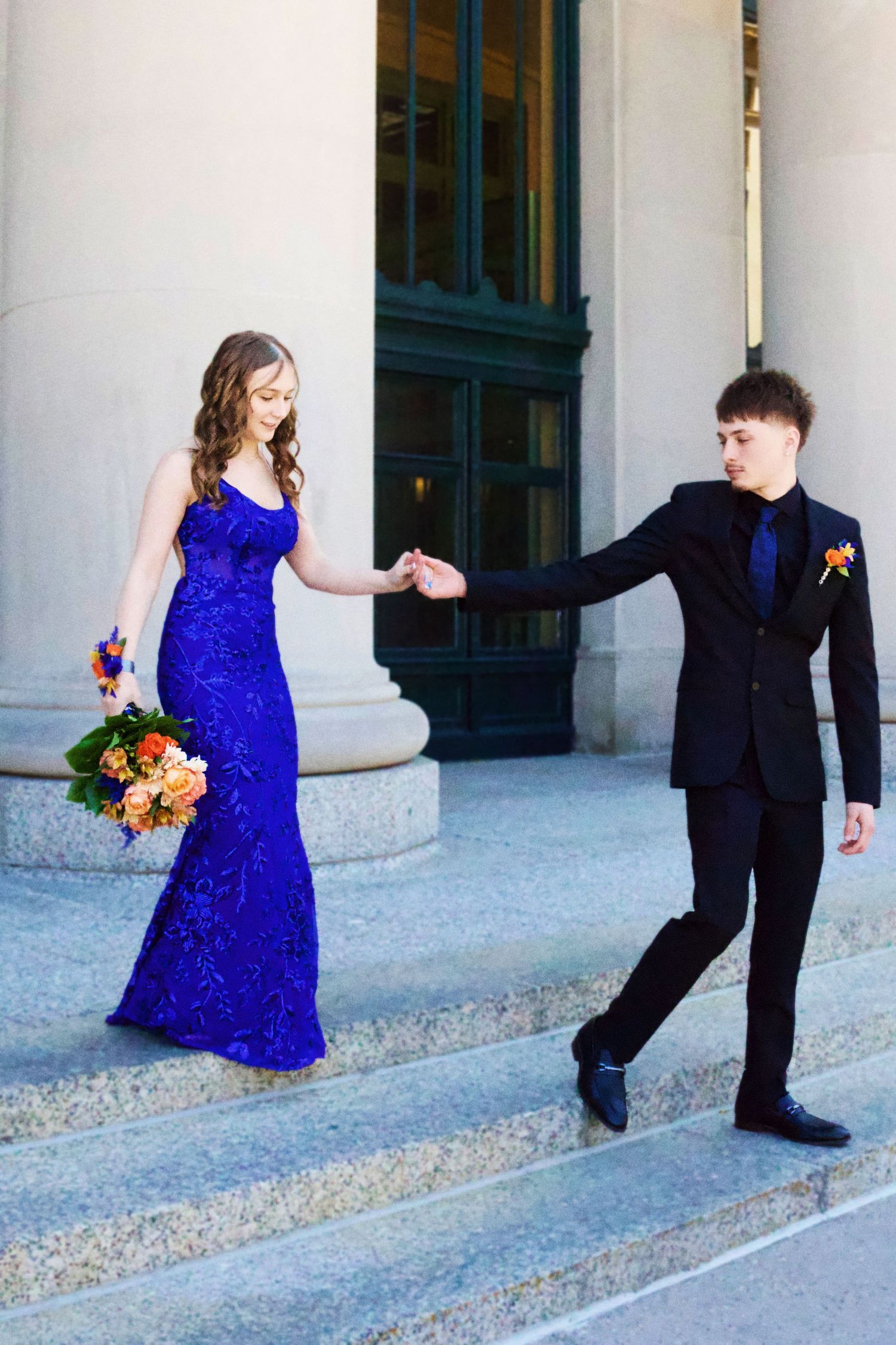 A couple in formal wear dances outdoors, the woman in a blue sequin gown, the man in a black suit.